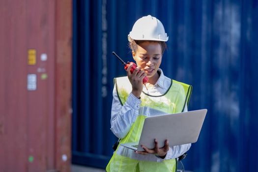 African American cargo container worker or engineer use walkie talkie to communicate other co-worker and hold laptop in workplace area.