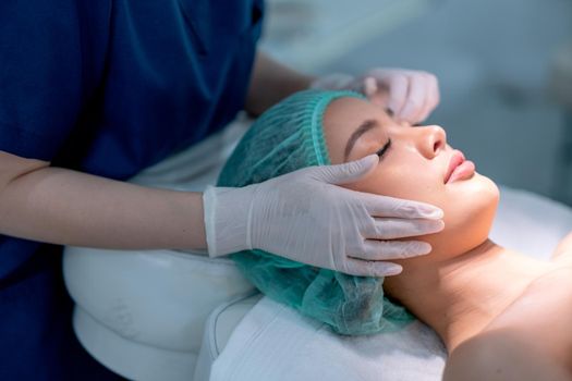 Beauty clinic hands touch on face of customer woman who lie on bed with light during process of treatment.