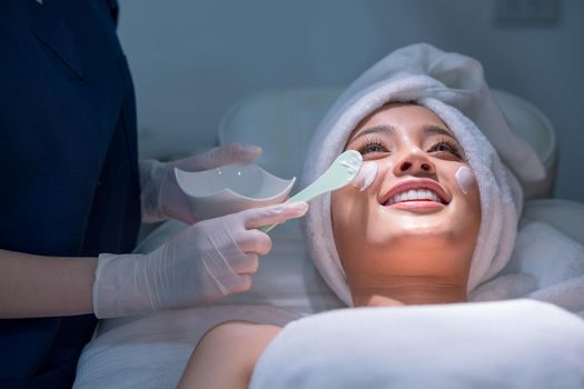 Close up hands hold paddle apply beauty cream on face of beautiful woman during process in beauty clinic or salon.