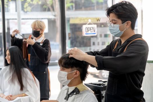 Beauty salon Asian barber man with hygiene mask work with hair of customer in the shop with other co-worker also take care hair style of woman. Beauty business for good appearance concept.