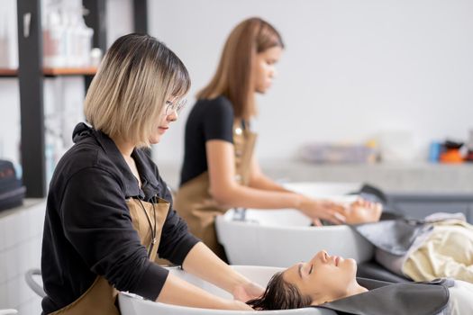 Asian barber girl is washing beautiful customer hair in beauty salon shop with other staff also work the same workplace. Beauty business for good appearance and healthcare concept.
