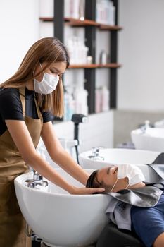 Asian beauty salon barber girl with hygiene mask wash man hair in workplace of the shop. Concept of beauty care business for good appearance of people.