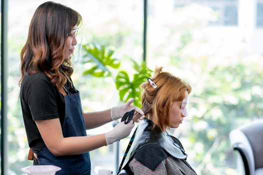 Beauty salon barber woman process of dyed Caucasian woman hair in the shop, both of them wear face shield during stay together.