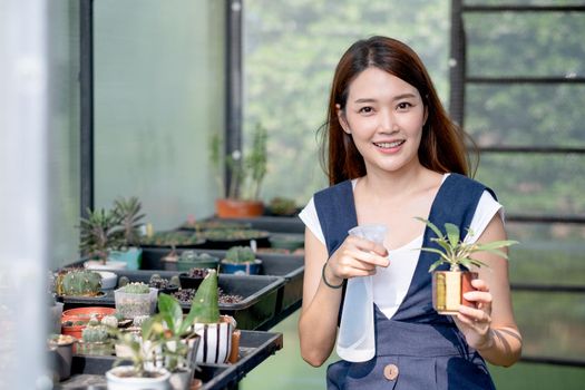 Beautiful Asian girl hold foggy bottle for spraying water to plant in small pot and sit in green garden in her house. Sustainable with grow cactus and other plant concept.