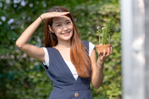 Beautiful Asian girl hold pot of plant with hand shading of light and stand near green house and smiling. Sustainable with small business relate to plant concept.