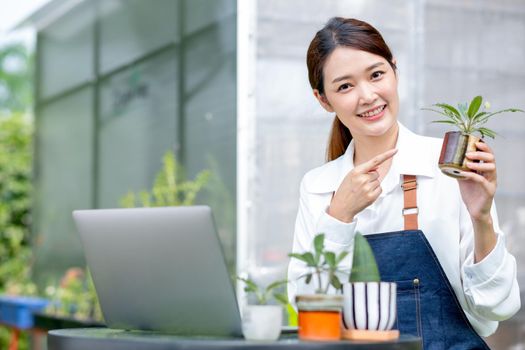 Beautiful Asian girl hold pot of plant and look at camera also pointing to pot stay with laptop on table in front green garden of her house.