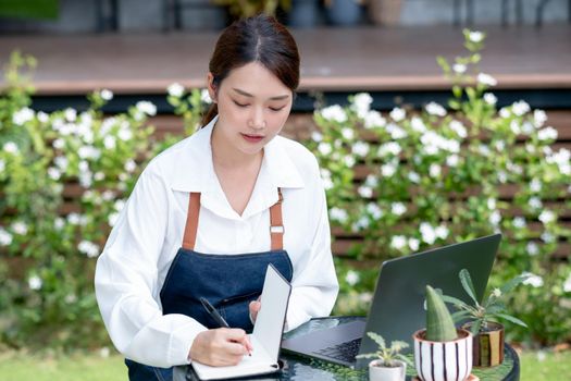 Beautiful Asian woman record the data into notebook in front of laptop and stay in home garden. Sustainable with small business relate to grow plant concept.