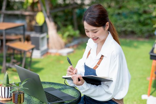 Beautiful Asian woman record the data into notebook in front of laptop and stay in home garden with happy emotion.