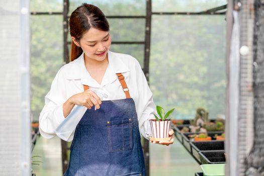 Beautiful Asian girl hold foggy bottle and spray water to plant in small pot and sit in green garden in her house. Sustainable with small business relate to grow plant concept.