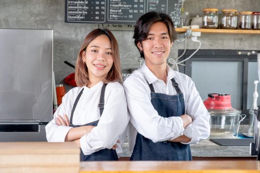 Two Asian barista or coffee maker man and woman stand with arm-crossed or confidence action also look to camera and smile in café shop. Concept of happy working with small business together.