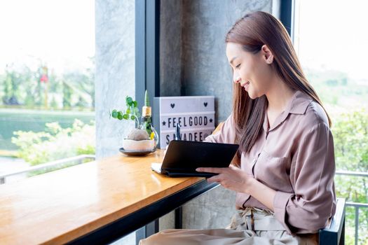 Beautiful Asian woman write some data on notebook and sit in corner of coffee shop with day light. She is smiling with happy emotion and think about her small business.