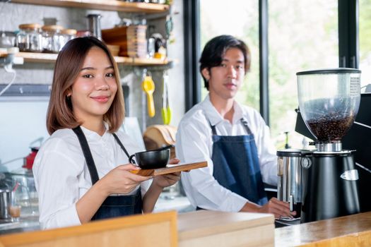 Barista woman or coffee maker hold plate with cup of coffee and look at camera in coffee shop and smiling. Concept of happy working with small business and sustainable together.