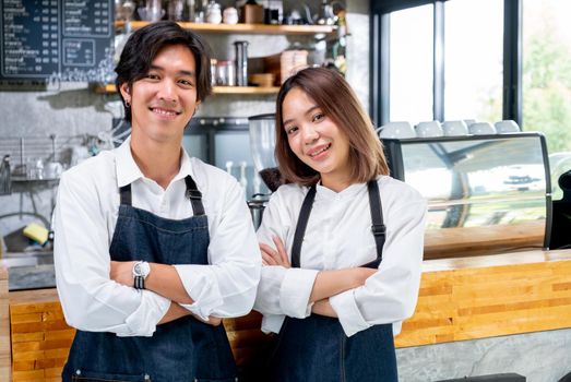 Two Asian barista or coffee maker man and woman stand with arm-crossed or confidence action also look to camera and smile in coffee shop. Concept of happy working with small business together.