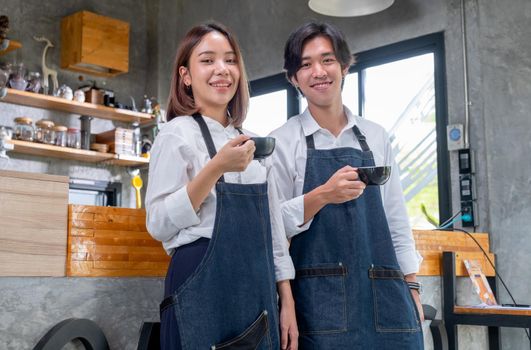 Two barista man and woman or coffee makers hold a cup and stand in front of counter in coffee shop with looking at camera and smile. Concept of happy working with small business and sustainable.