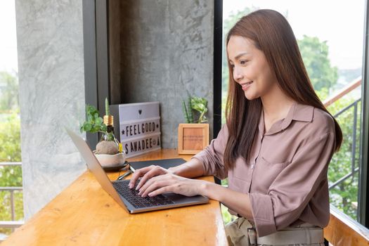 Beautiful Asian woman work with laptop and sit in corner of coffee shop with day light. She is smiling with happy emotion and think about her small business and sustainable activity.