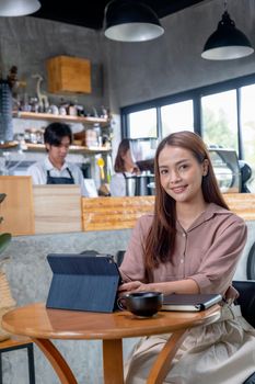 Beautiful Asian woman work with tablet in coffee shop, she also look at camera and two barista or coffee maker work together in the back. Concept of small business and support by customer.