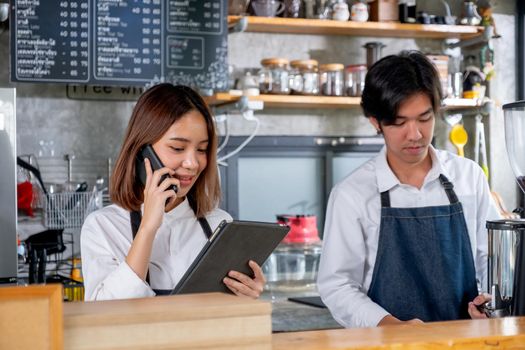Barista girl or coffee maker receive phone call and record order and stand beside of co-worker man in coffee shop. Concept of happy working with small business and sustainable together.
