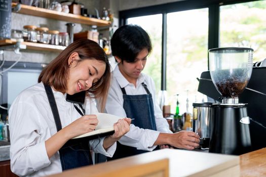 Barista girl or coffee maker answer mobile phone also record the order detail into notebook while her co-worker is working with machine. Concept of good teamwork support small business system.