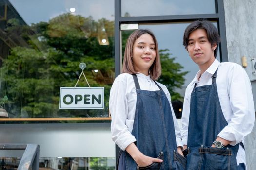 Two Asian barista or coffee maker man and woman stand with confidence action also look at camera and smile in front of coffee shop with day light.