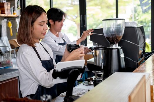 Barista girl or coffee maker woman use towel clean cup of coffee while her co-worker work with machine. Concept of happy working with small business and sustainable.