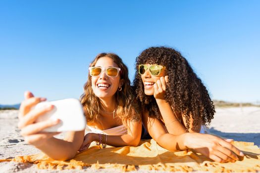 Beautiful young woman holding smartphone taking a picture with her curly black Hispanic girlfriend lying on beach with sunglasses smiling looking at the camera. Summer is good for travel and tourism