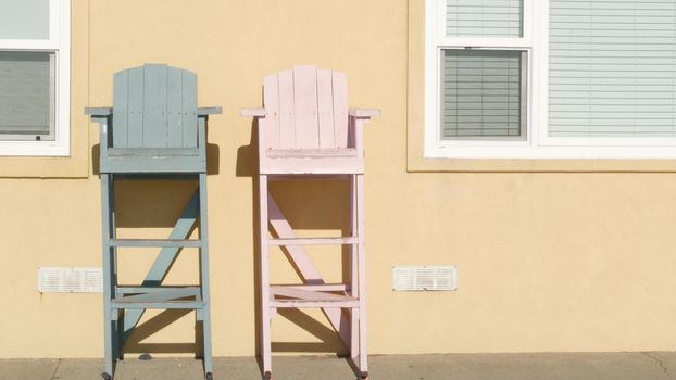 Two vintage wooden lifeguard chairs by yellow wall, California USA. Empty life guard retro high seat by ocean sea beach. Minimal simple trendy pop atmosphere, summertime aesthetic. Los Angeles vibes.