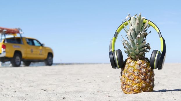 Funny pineapple in headphones, sandy ocean beach, yellow lifeguard car, California USA, Pacific coast. Tropical summer exotic fruit enjoying vacations and music in paradise. Ananas sunbathing on shore