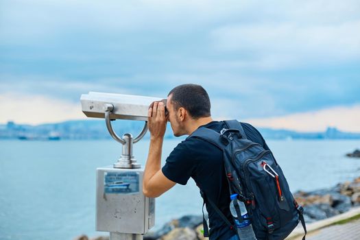 A tourist guy with a backpack on his back looks through the sightseeing binoculars on the observation deck.