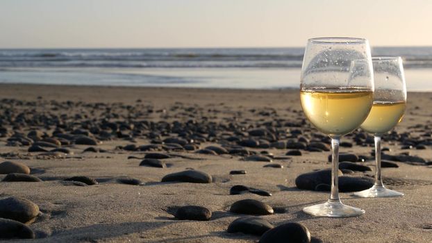Two wineglasses on sandy ocean beach. Couple of glasses and pebbles, white wine for romantic date near sea water. Cozy beachfront sunset near tide waves. Seamless looped cinemagraph. California, USA.