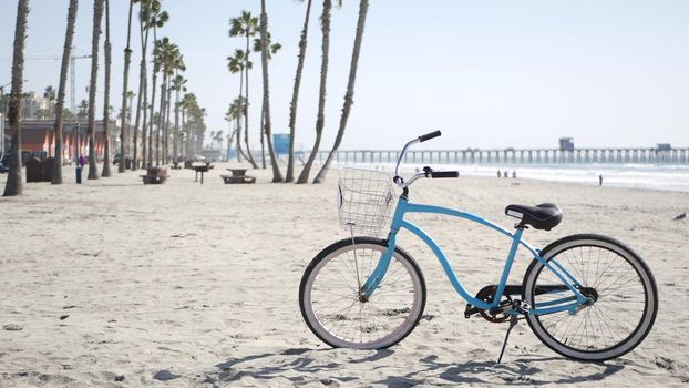 Blue bicycle, cruiser bike by sandy ocean beach, pacific coast near Oceanside pier, California USA. Summertime vacations, sea shore. Vintage cycle, tropical palm trees, lifeguard tower watchtower hut