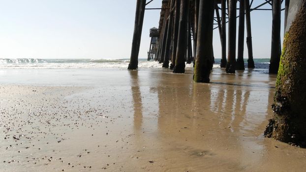 Wooden piles under boardwalk, old pier in Oceanside, California coast USA. Pilings, pylons or pillars below retro vintage bridge, waterfront promenade. Ocean waves, sea water tide and sand beach.