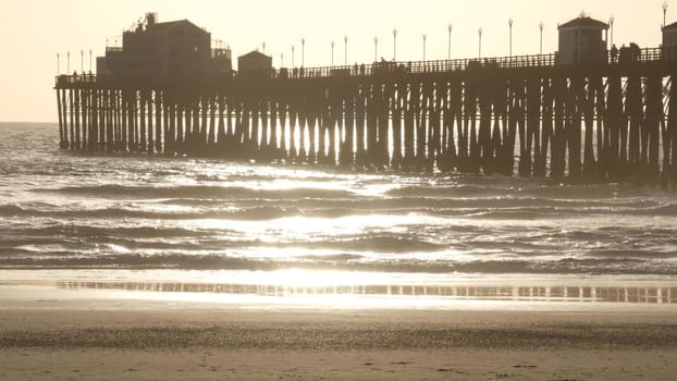 Wooden pier on piles, silhouette at sunset, California USA, Oceanside. Waterfront resort, pacific ocean tide, tropical beach. Summertime coastline vacations atmosphere. Sunny sea waves at sundown.