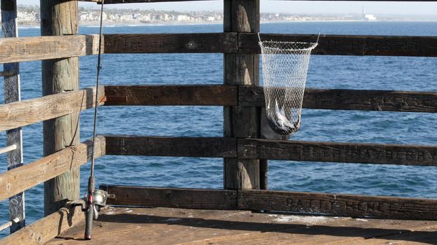 Saltwater angling, wooden pier boardwalk, fishing accessory, tackle or gear. Oceanside California USA. Sea salt water, ocean seascape. Rod or spinning, fisherman catch, fresh alive fish in net basket.