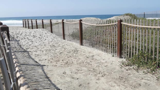 Pacific ocean coast, greenery and wooden picket fence on sea shore. Blue water waves on sunny summer beach, Encinitas shoreline, California USA. Coastline near Los Angeles. Coastal access entrance.