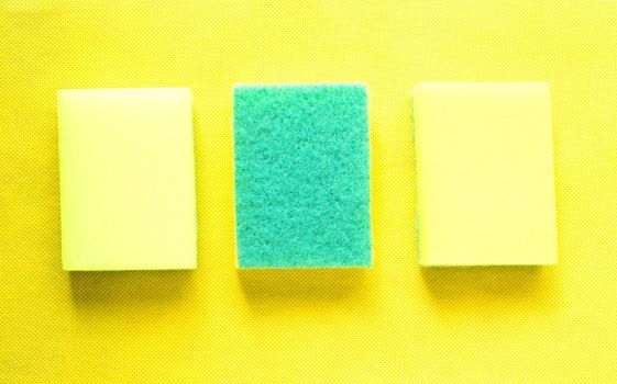 Two bright yellow sponges for cleaning surfaces and one upside-down to remove stubborn dirt on a yellow background.