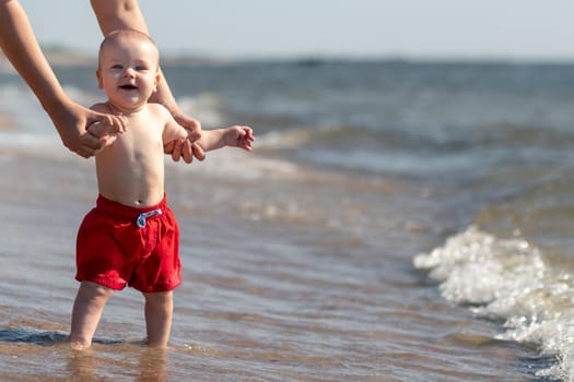 Funny baby boy for the first time by the sea trying to walk in the water with the help of her mom