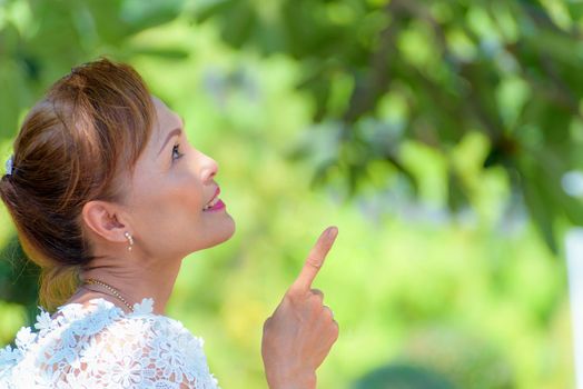 Side face of a beautiful woman healthy middle-aged Asian people dress in Thai style clothes, Portrait old lady relaxing outdoors pointing finger looking up in the park, copy space on nature background