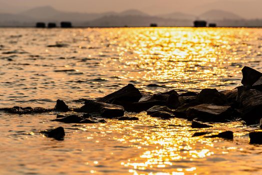 Beautiful nature landscape of bright golden sunlight reflects the yellow glow on the ripples pile of rock in the water of a tropical lake mountain background at Krasiao Dam, Suphan Buri, Thailand