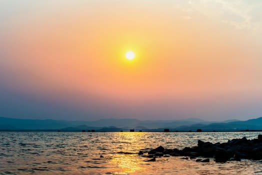 Beautiful nature landscape yellow sun in the orange sky lake reflecting the golden sunlight on the water surface at sunset, fish farm cages mountain background at Krasiao Dam, Suphan Buri, Thailand