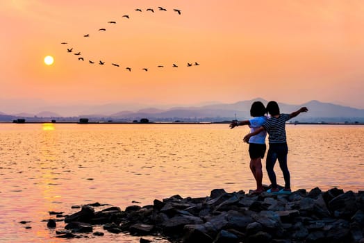 Mother and daughter were happy by the water spread out his arms to fly a flock of birds flying in a row on the sky sun at sunset beautiful nature landscape background, Krasiao Dam Suphan Buri Thailand