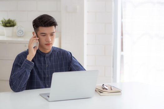 Young asian man talking phone and work from home with laptop computer, freelance using notebook to internet online and speak on smartphone, domestic life, business and communication concept.