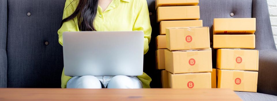 Young asian woman is merchant using laptop computer on sofa checking order of customer with purchase online shopping, business SME or startup, entrepreneur or seller and service delivery product.