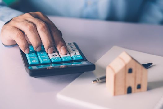 Hand of businessman planning and calculate expense and mortgage with calculator and home on desk, insurance and budget of residential, examining of loan and residence, business and property concept.