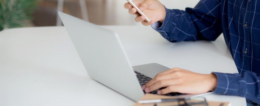 Closeup hand of young man working laptop computer and reading smartphone on internet online on desk at home, freelance male using phone with social media, business and communication concept.