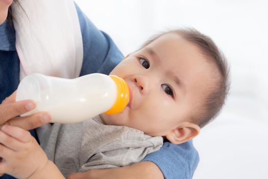 Young asian mother embracing and feeding little baby girl with bottle of milk at home, newborn innocence drinking with mom satisfied, relationship and bonding of mum and child, family concept.
