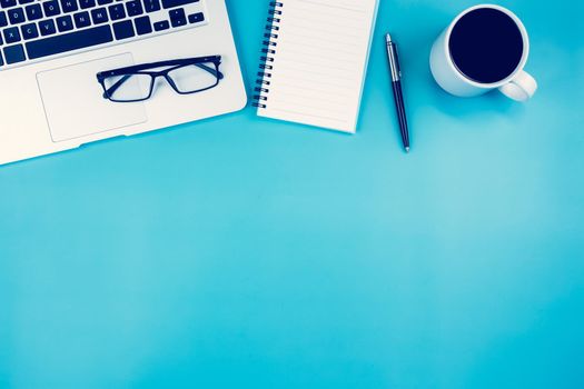 Flat lay of laptop computer and cup of coffee on desk in office, workplace and notebook and glasses and book on blue background, workspace and copy space, top view, business and communication concept.