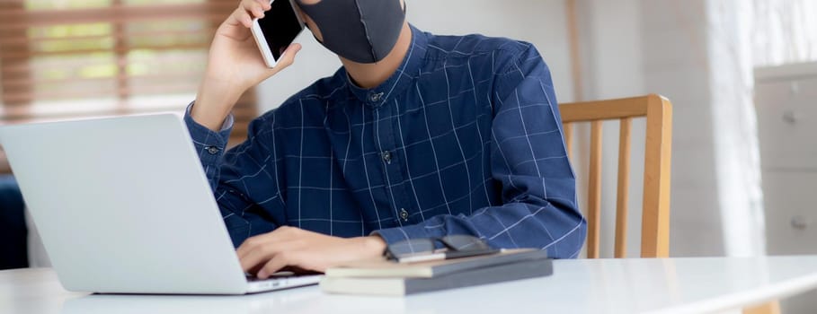 Young asian business man in face mask talking smartphone and work on laptop computer for protect covid-19, businessman quarantine work from home with notebook, social distancing and stay home.