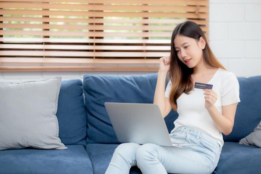 Young asian woman sit on sofa using laptop computer shopping online with credit card buying to internet, happy girl payment with e-business on couch, purchase and payment, business concept.