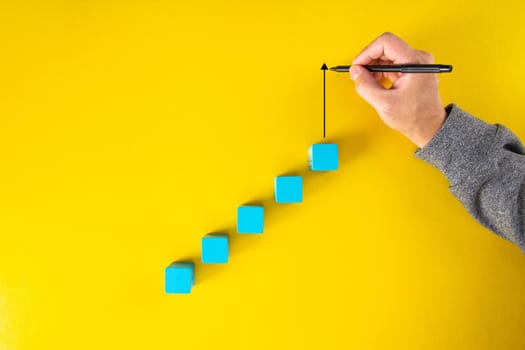 Man hand drawing an upward pointing arrow on top of growing graph made of wooden block. Over yellow background