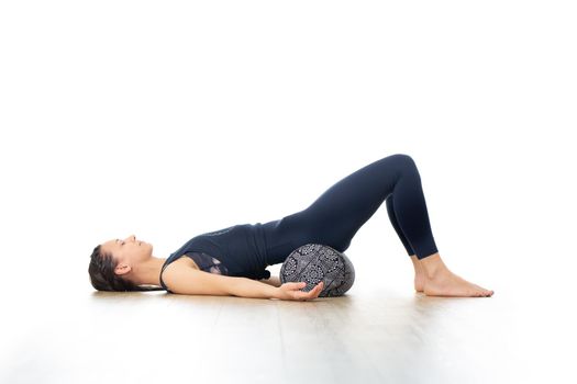 Restorative yoga with a bolster. Young sporty attractive woman in bright white yoga studio, lying on bolster cushion, stretching and relaxing during restorative yoga. Healthy active lifestyle.
